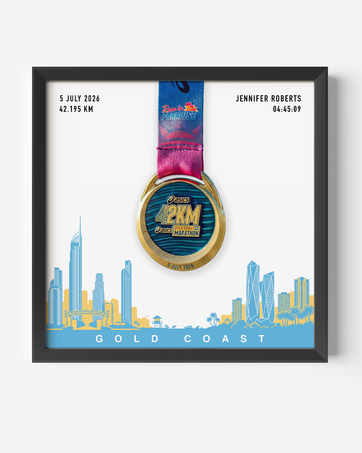 A framed personalized Gold Coast marathon medal print featuring a gold and blue 2KM medal with a pink and blue ribbon, displayed on a white background with a blue and yellow coastal city skyline and the text 'GOLD COAST'. Personalized details for Jennifer Roberts are shown on the print.