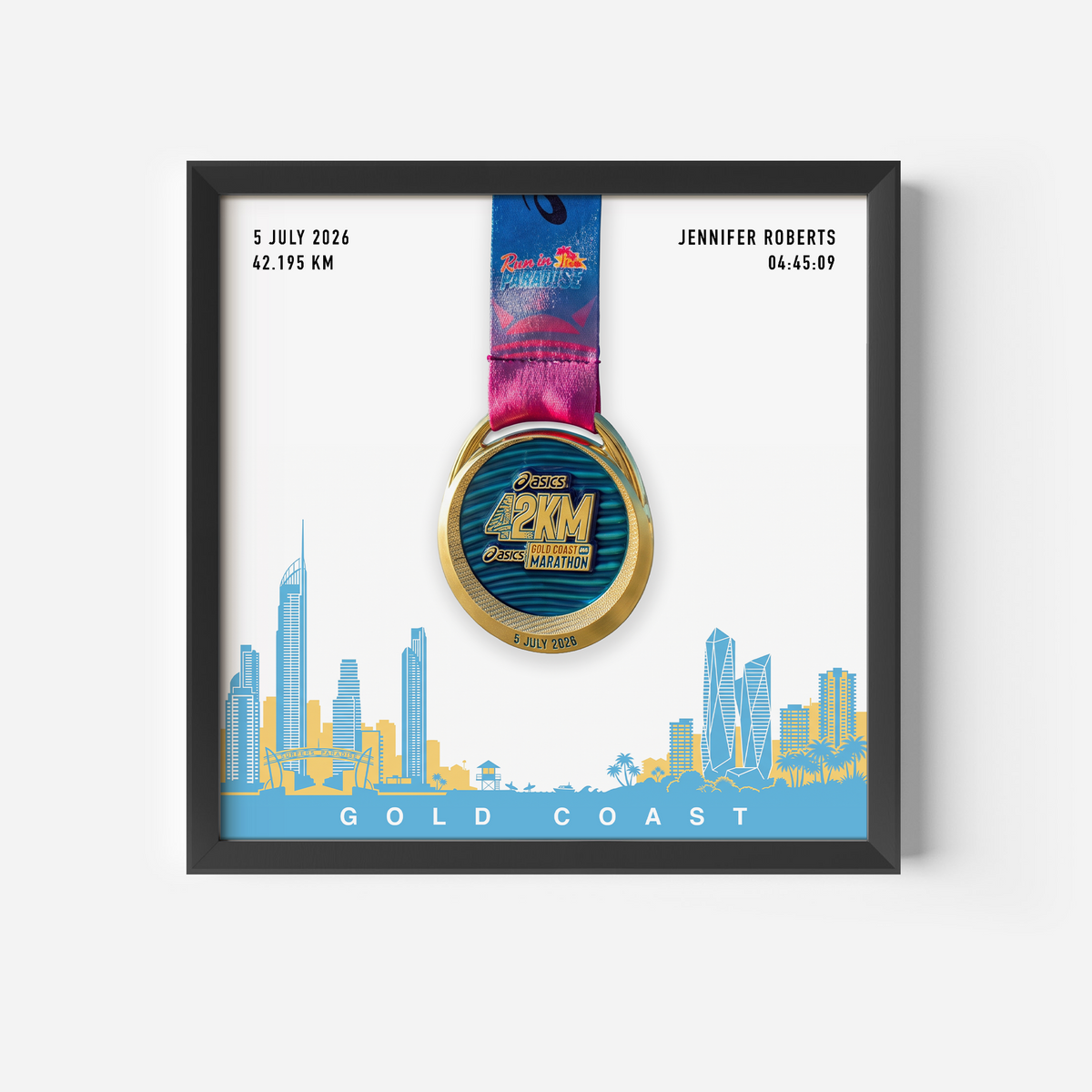 A framed personalized Gold Coast marathon medal print featuring a gold and blue 2KM medal with a pink and blue ribbon, displayed on a white background with a blue and yellow coastal city skyline and the text 'GOLD COAST'. Personalized details for Jennifer Roberts are shown on the print.