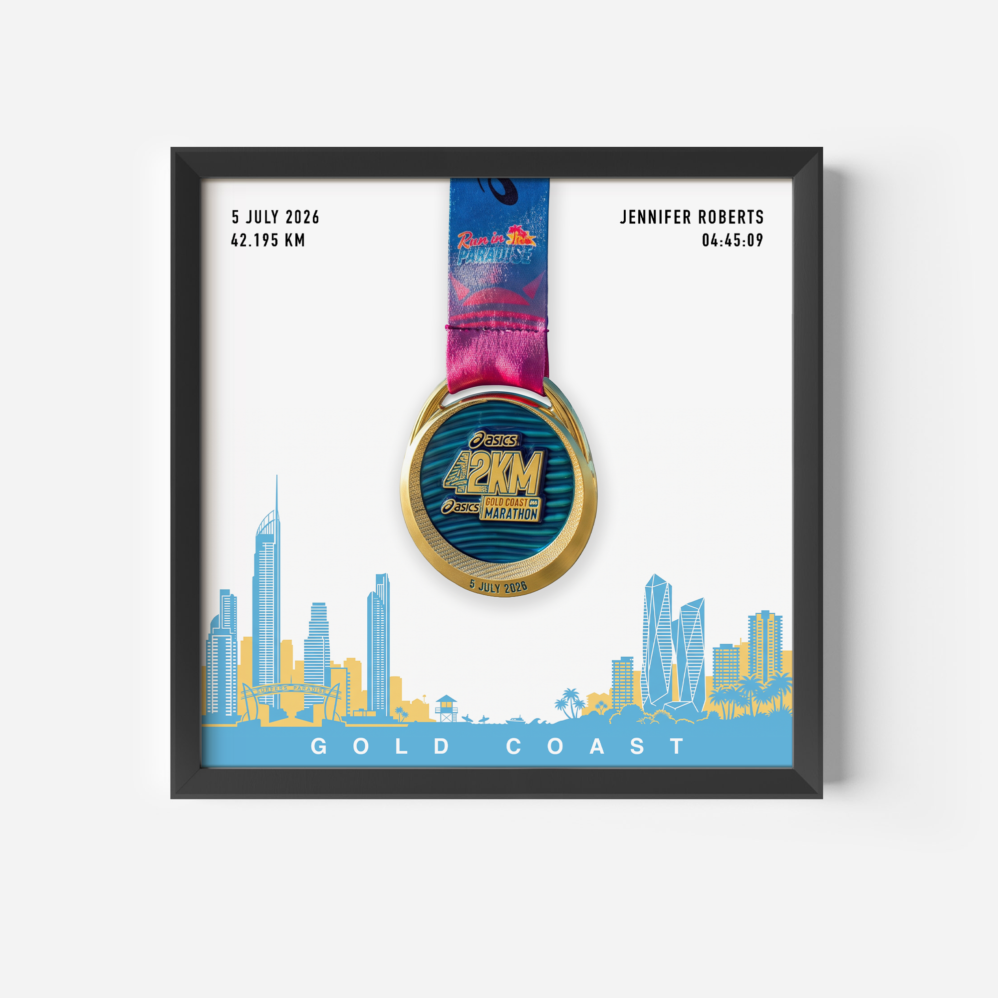 A framed personalized Gold Coast marathon medal print featuring a gold and blue 2KM medal with a pink and blue ribbon, displayed on a white background with a blue and yellow coastal city skyline and the text 'GOLD COAST'. Personalized details for Jennifer Roberts are shown on the print.